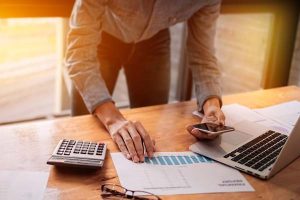 Man bends over table paying bills with laptop, calculator, and smartphone. Glasses and a spreadsheet sit on desk.